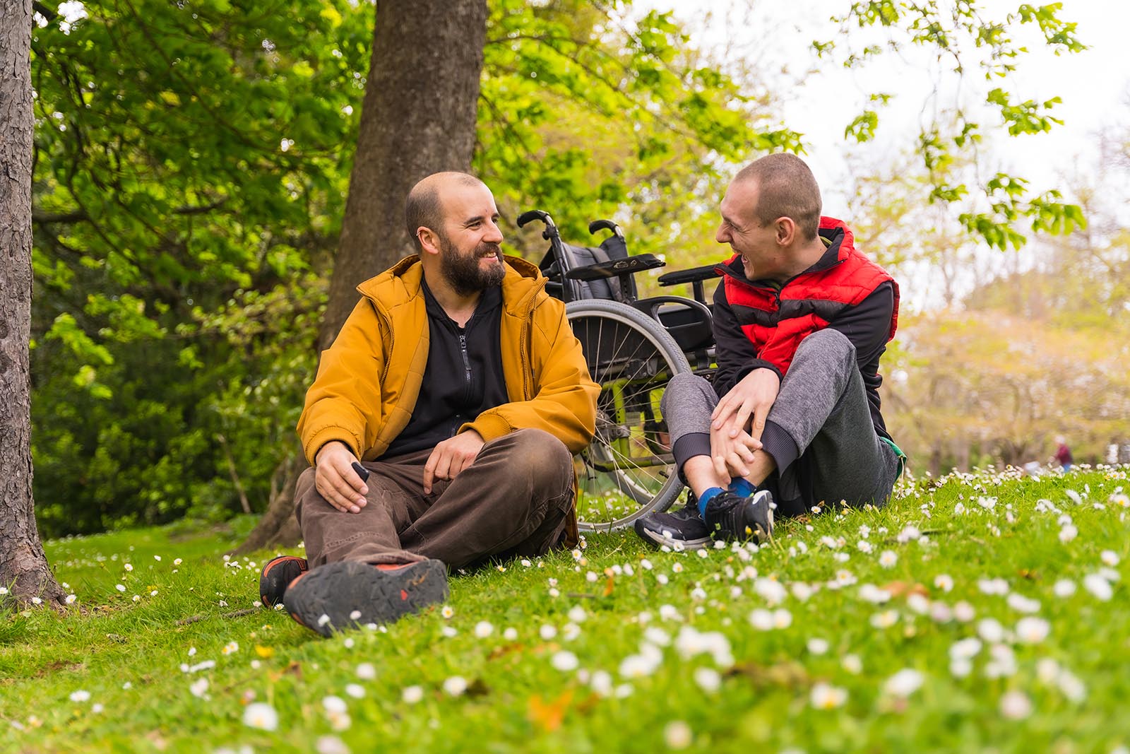 Zwei Männer sitzen im Gras, im Hintergrund ist ein Rollstuhl zu sehen.