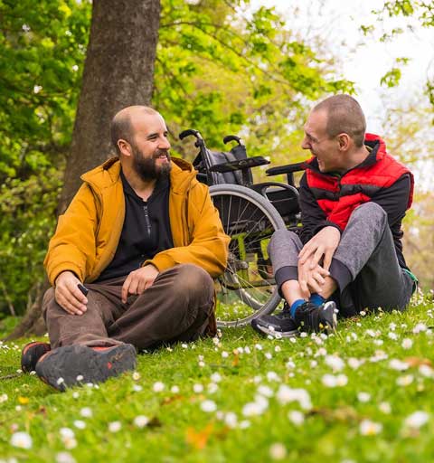 Zwei Männer sitzen im Gras, im Hintergrund ist ein Rollstuhl zu sehen.